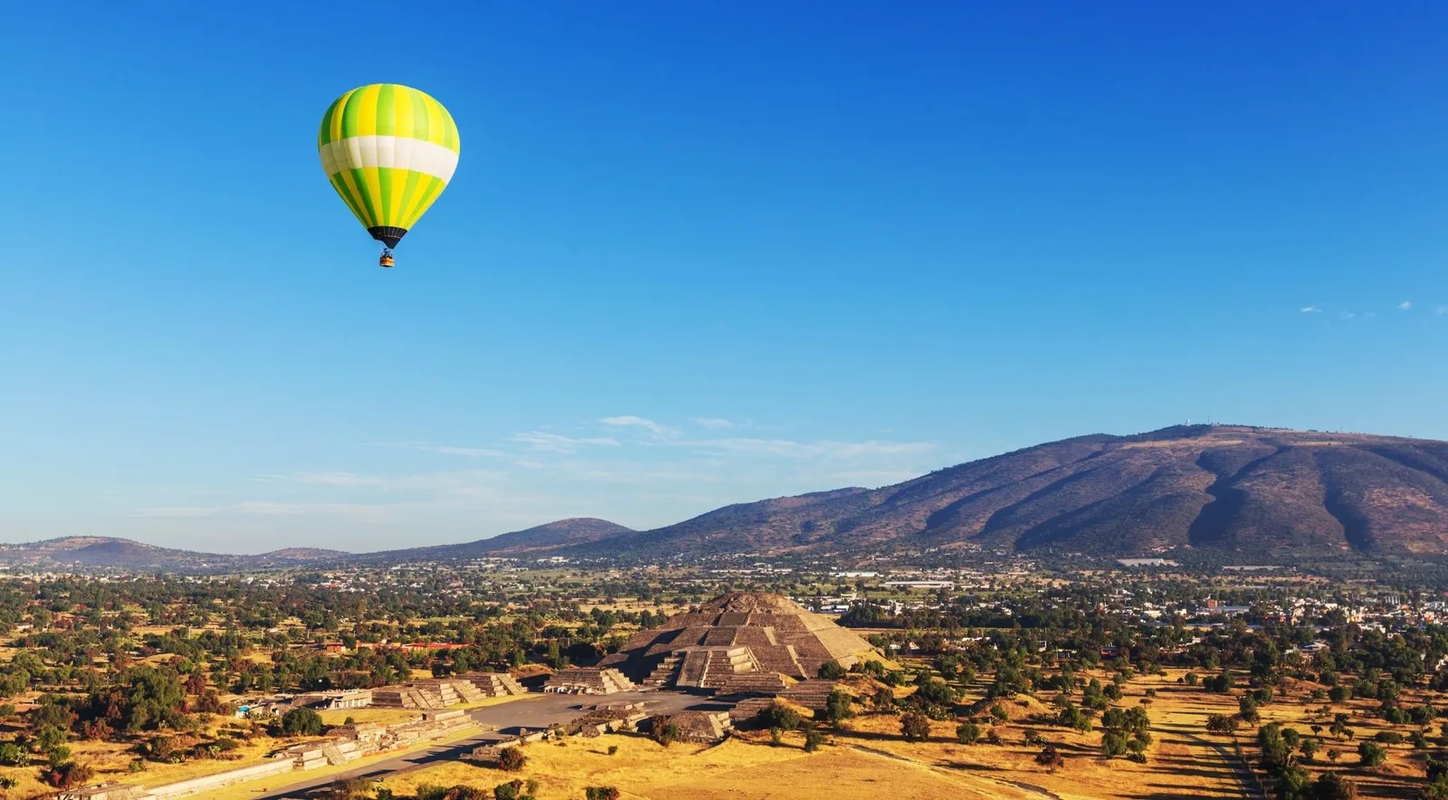 A green, yellow and white hot-air balloon in a clear blue sky above the Pyramid of the Moon, with dry fields, trees and a long mountain behind