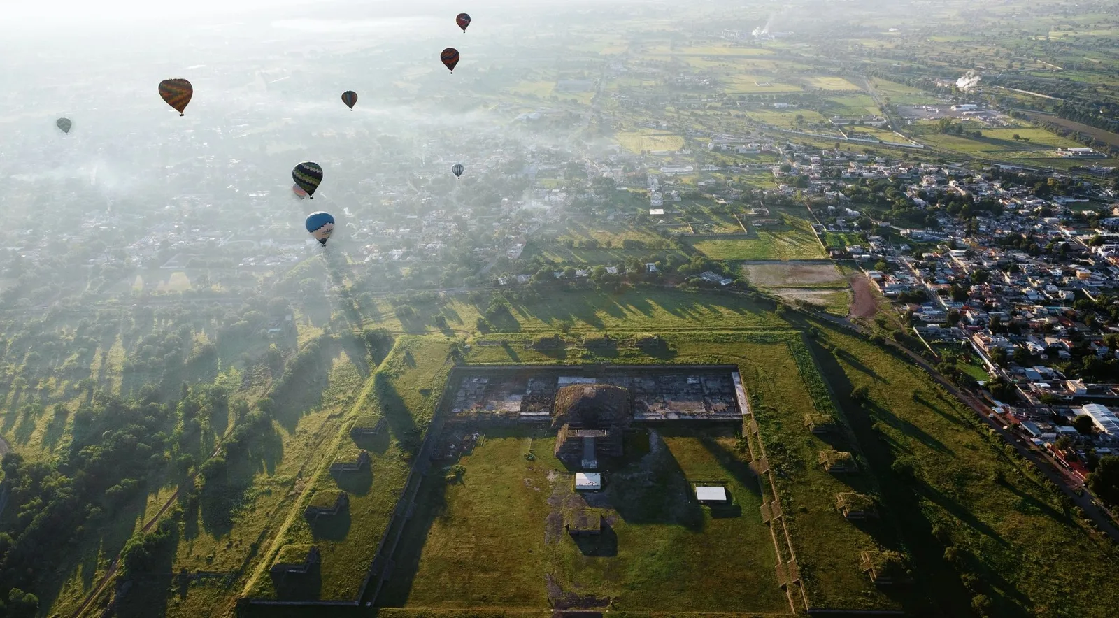 Aerial view of balloons drifting through morning haze over the square walled enclosure of the Temple of Quetzalcoatl, with a town and green fields beyond