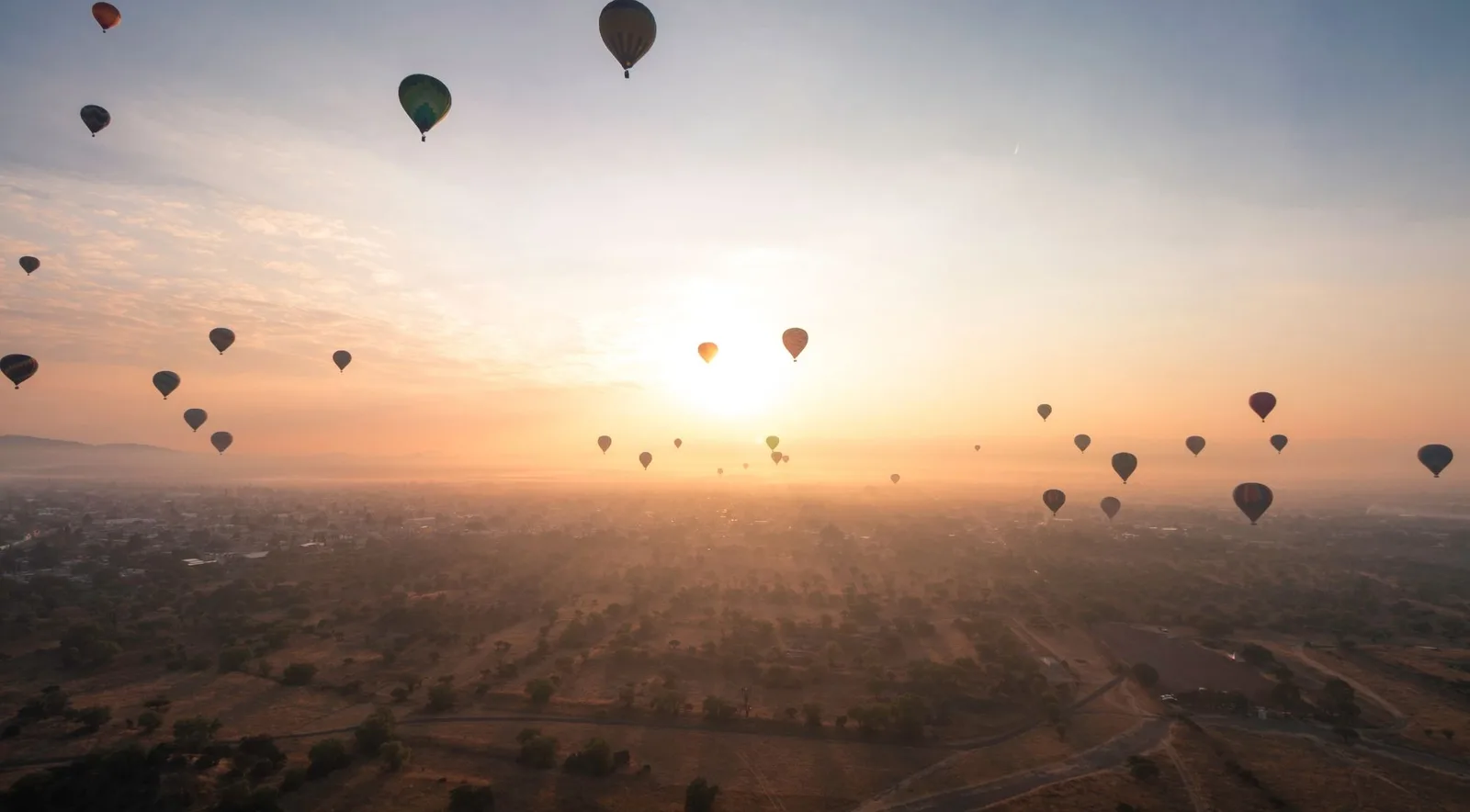 Dozens of hot-air balloons silhouetted against the rising sun over the hazy Teotihuacán valley
