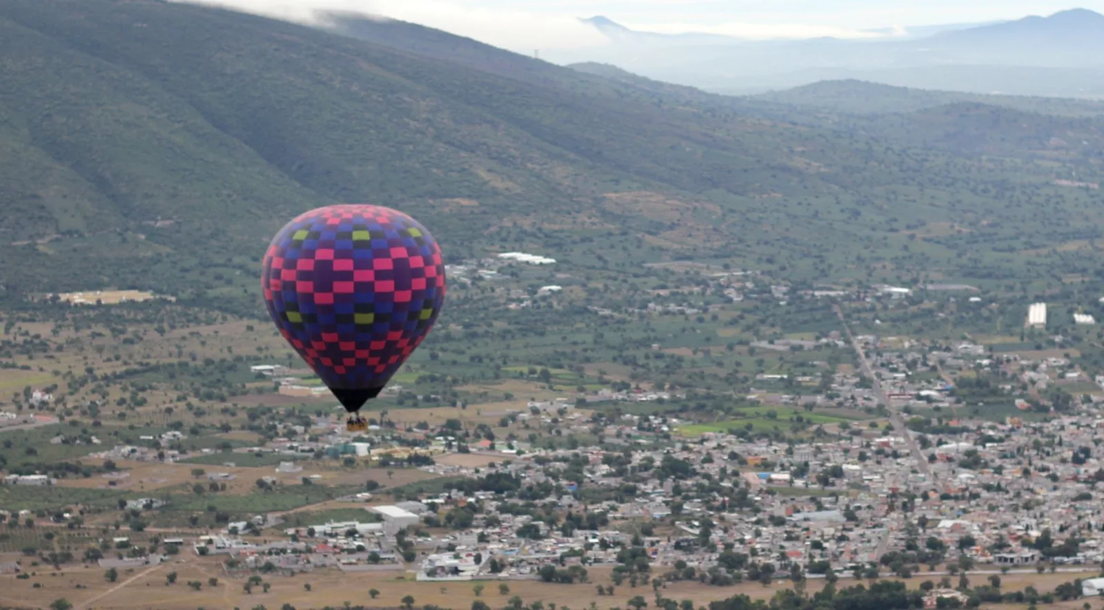 A pink, purple and blue checked balloon flying over a small town and fields, with green hills behind