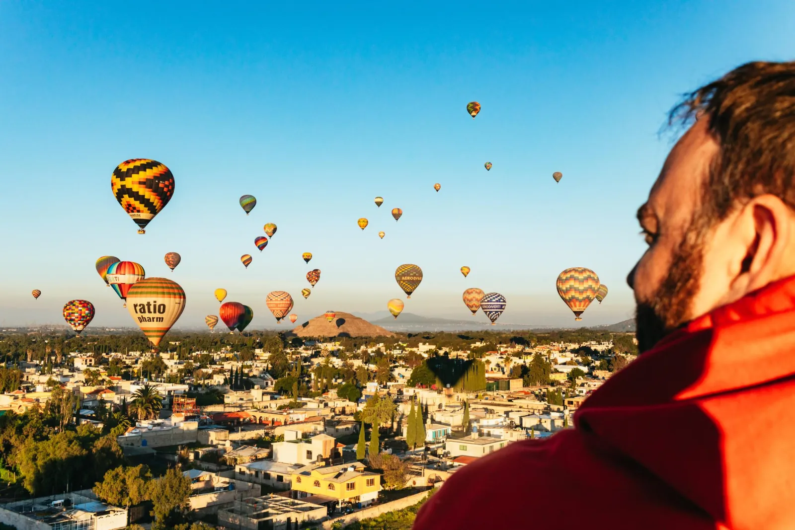 Passenger in a red jacket looks out at balloons over the rooftops of a town