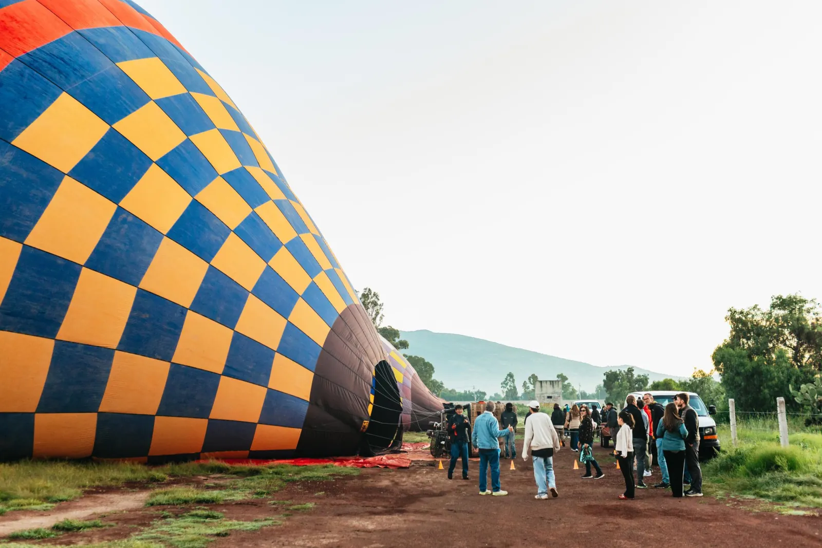 Passengers walking near a checkered balloon envelope laid out on a dirt launch field
