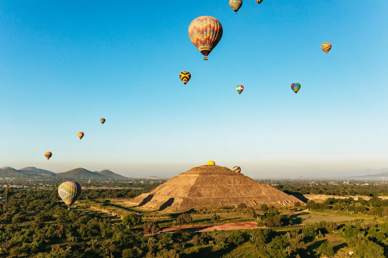 Large stepped pyramid among green fields with balloons floating in a clear sky