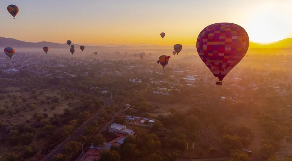 Dozens of hot-air balloons rising at sunrise over the Teotihuacán valley, with a red-checked balloon in the foreground