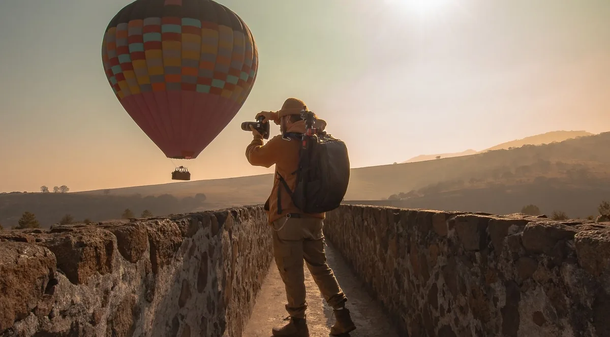 A photographer with a backpack and tripod stands on an old stone wall photographing a checkered hot air balloon in the hazy early sun over the Teotihuacán valley