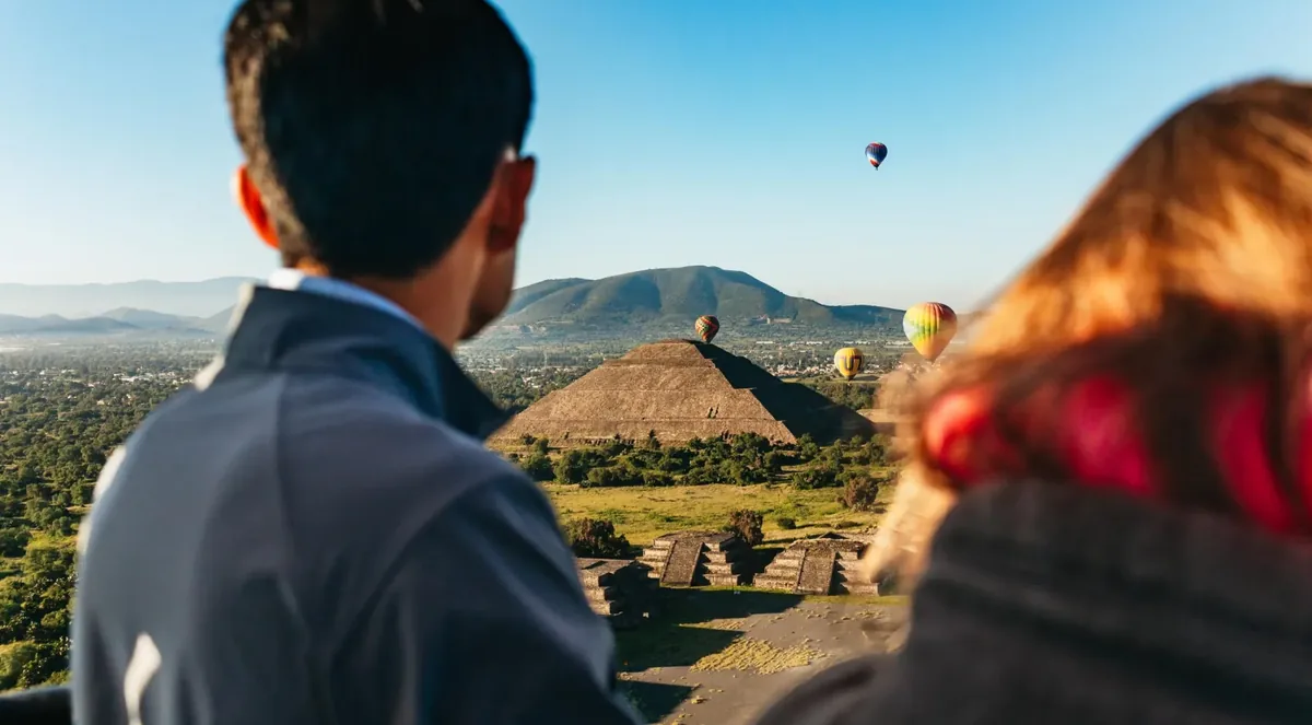 Two passengers seen from behind in a balloon basket, looking toward the Pyramid of the Sun with other balloons in a clear blue sky