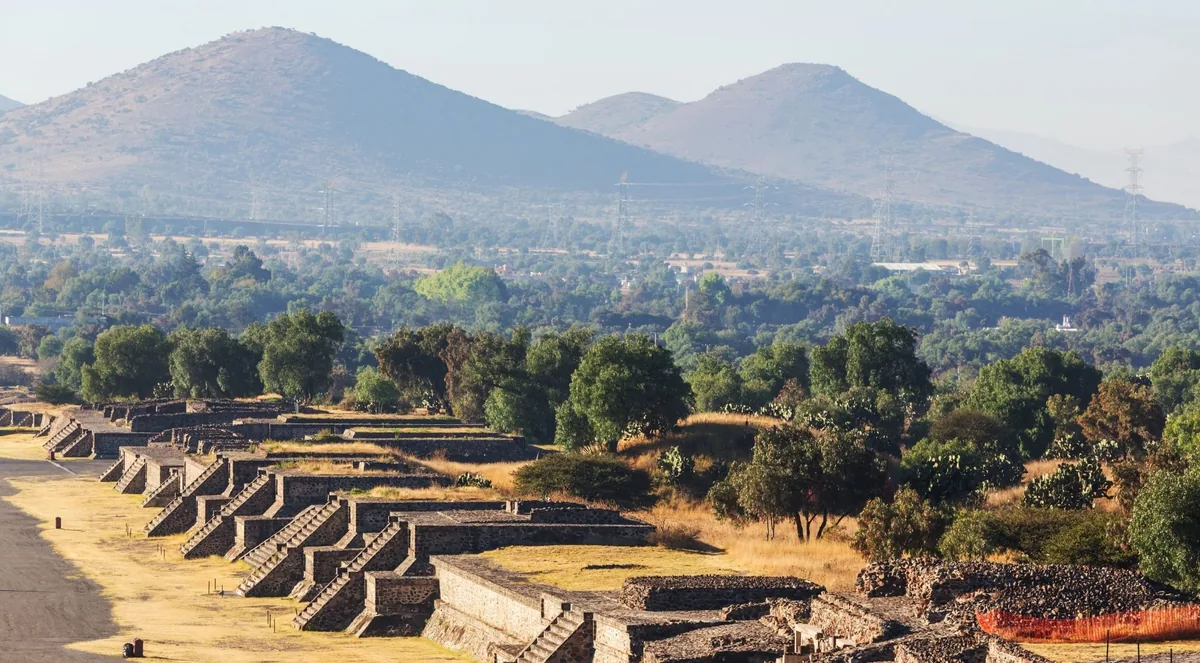 Rows of stepped stone platforms along the Avenue of the Dead at Teotihuacán, with trees and two hills behind in morning haze