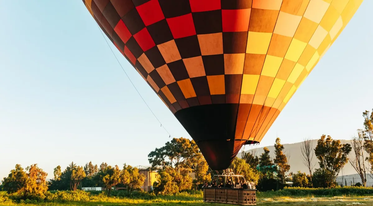 A red, orange and yellow checkered hot air balloon, fully inflated, with passengers in its basket on a grassy field at dawn, trees and a hill behind
