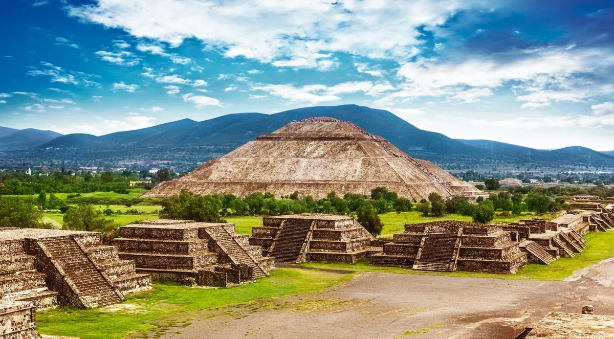 The Pyramid of the Sun at Teotihuacán seen from the Avenue of the Dead, with stepped stone platforms in the foreground and green hills under a blue sky