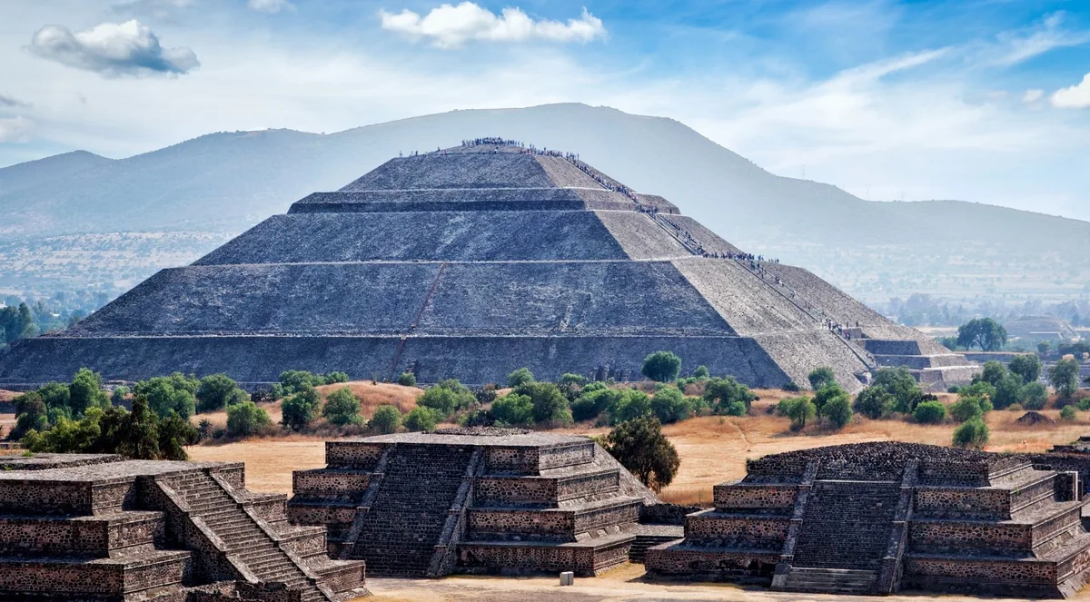 The stepped Pyramid of the Sun at Teotihuacán under a blue sky, with low stone platforms in the foreground and hills behind