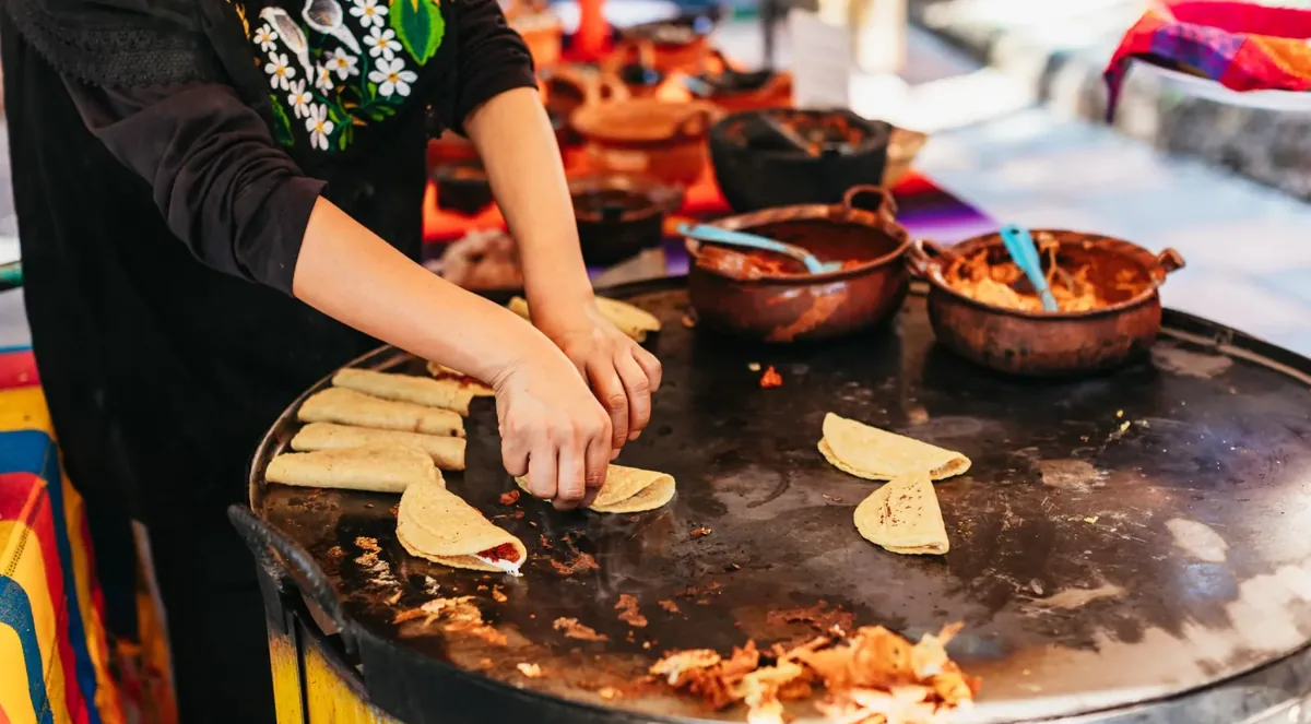 A cook in an embroidered black blouse folding quesadillas on a large round comal, with clay pots of fillings behind