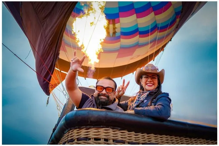 Couple waving from the basket under a burner flame with a colorful balloon overhead