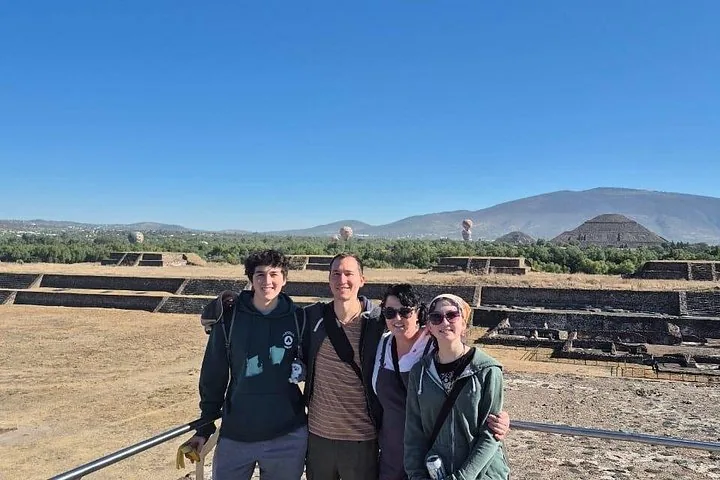Four people posing on a ruins terrace with a pyramid and distant balloons behind