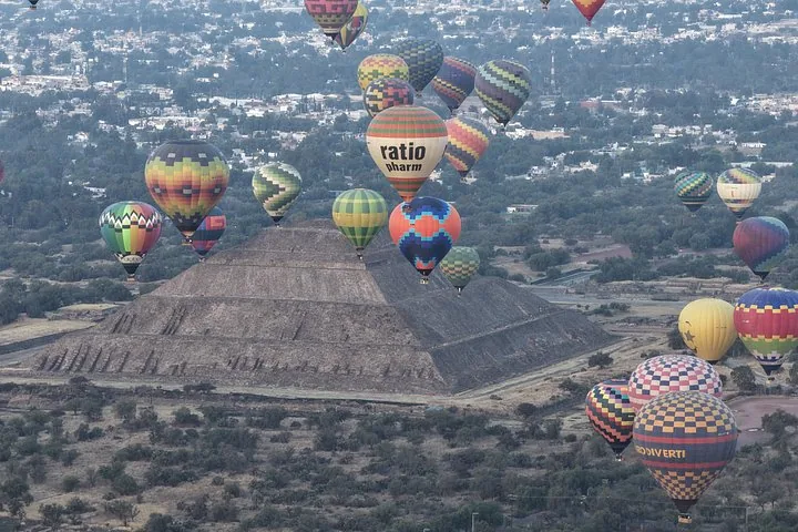 Many balloons rising around a stepped pyramid with the town of Teotihuacán beyond
