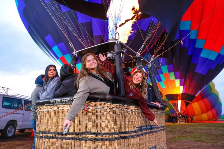 Passengers waving from a wicker basket while a nearby balloon inflates with flame