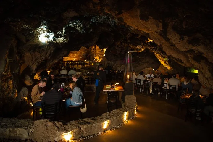 Diners at tables inside a lit natural cave restaurant with a curved stone wall