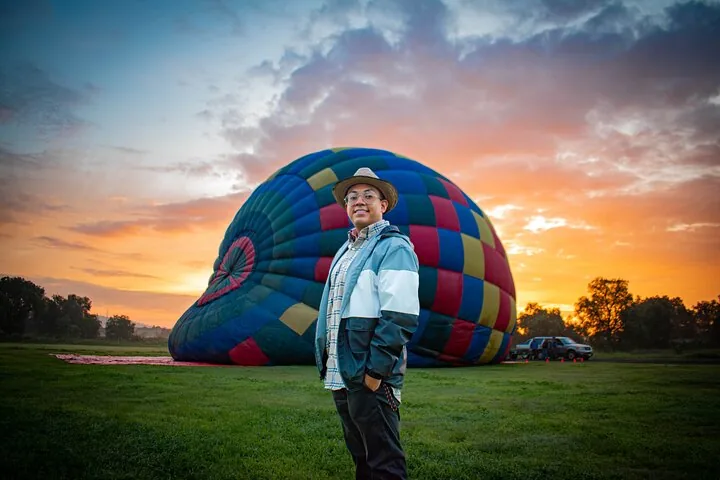 A man in a hat standing before a half-inflated balloon at orange sunrise