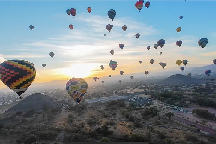 Dozens of balloons rising at sunrise above the valley, with a pyramid and hills below