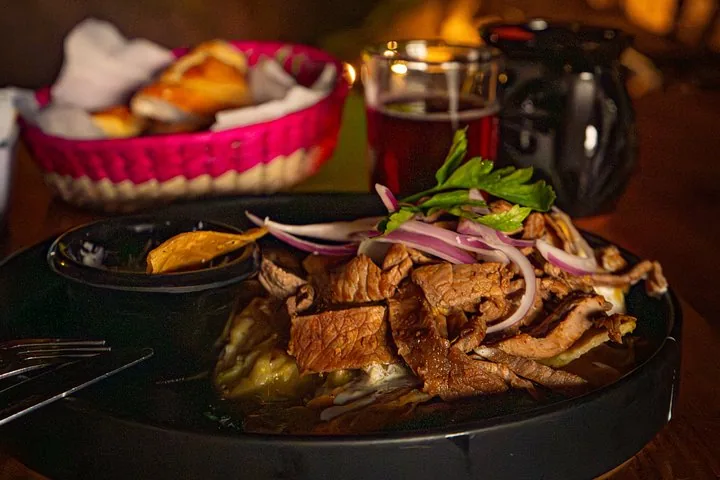 A plate of grilled meat with a bread basket and a drink on a dark table