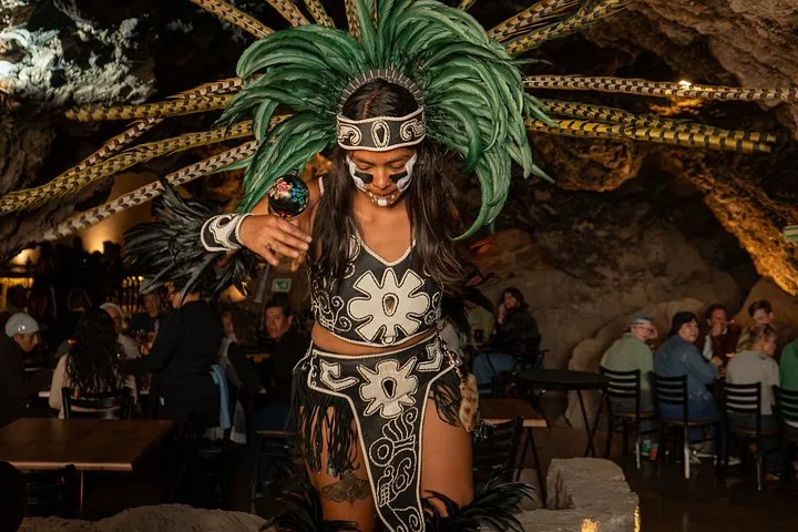 A performer in a green feather headdress dancing inside a cave restaurant with diners behind
