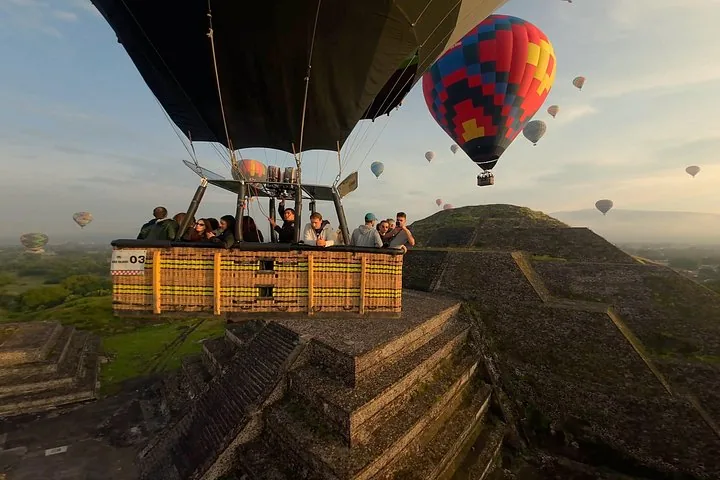 Basket of passengers flying close beside the stepped side of a pyramid with balloons around